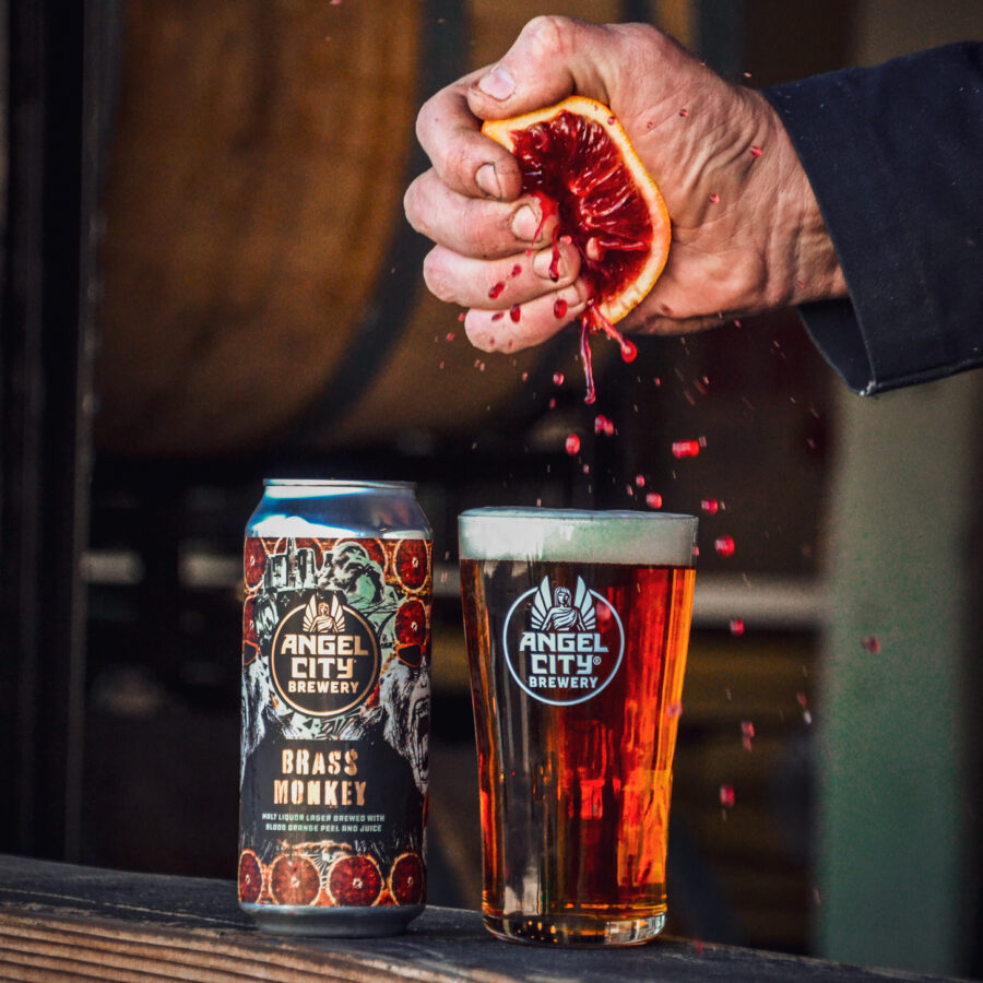Can of Brass Monkey Malt Liquor Lager next to a pint of the same beer, with a hand squeezing a blood orange into the beer.
